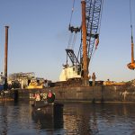 Crane Lowering Concrete Into Water & Men Watching From Rowboat Near Long Island, NY